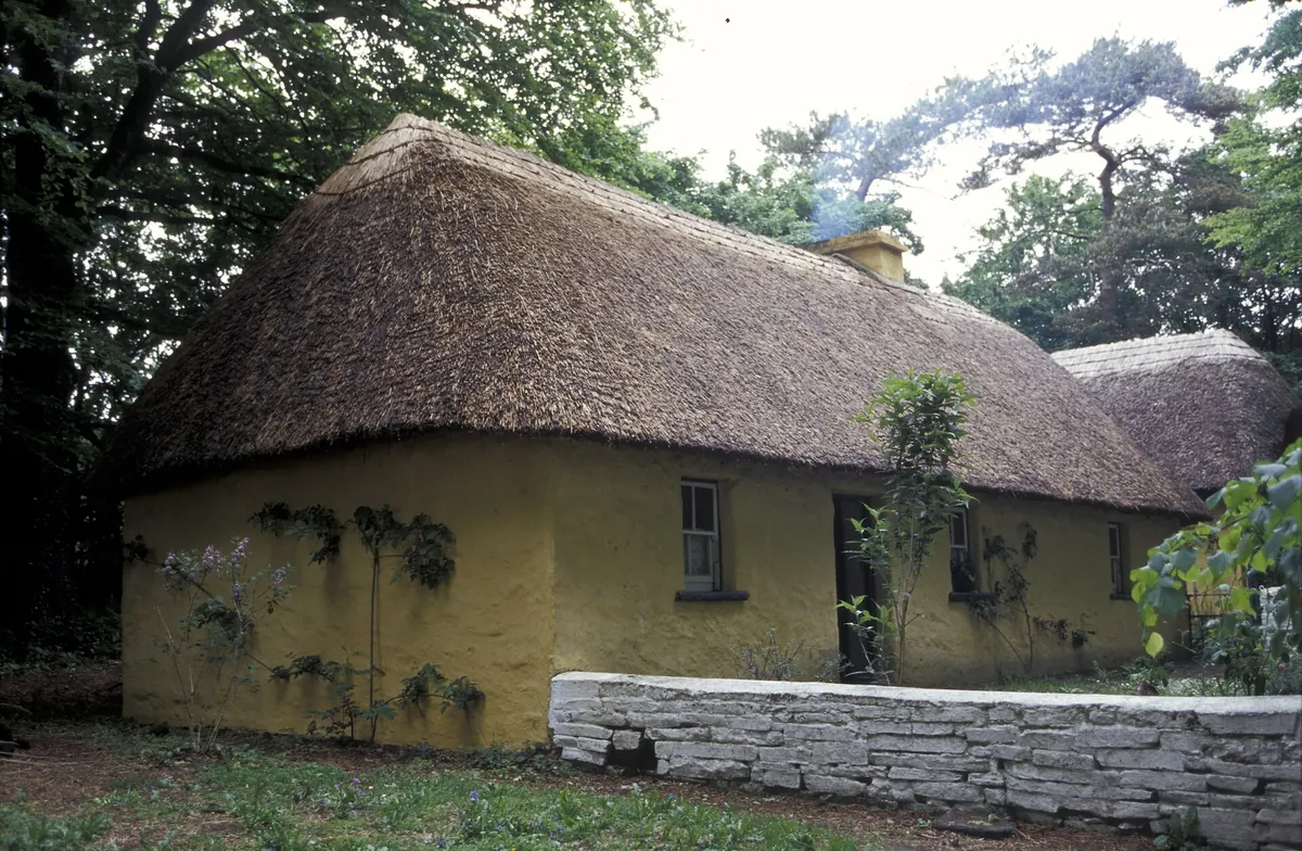 A yellow-walled thatched cottage in Bunratty Folk Park with a low stone wall in front and trees behind
