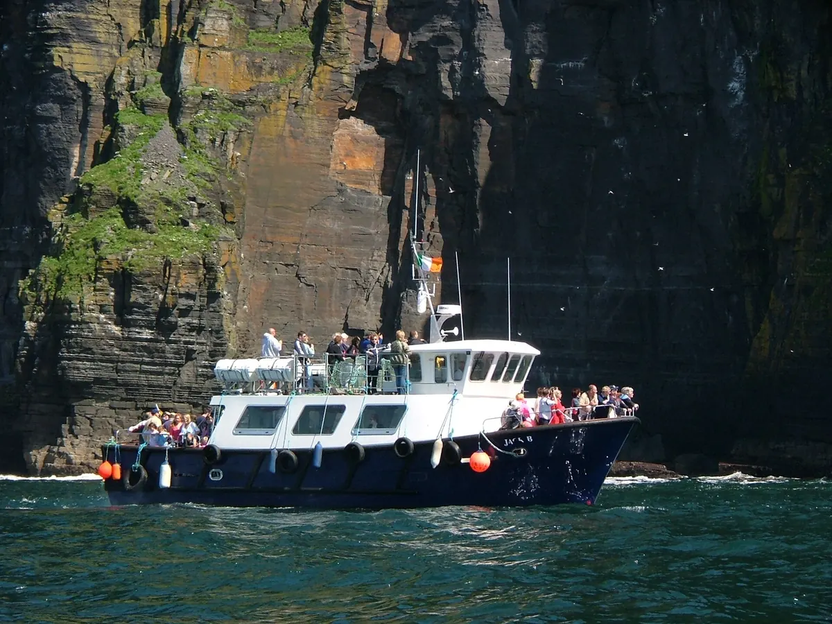 A white and navy passenger boat with people on deck at the foot of tall dark sea cliffs