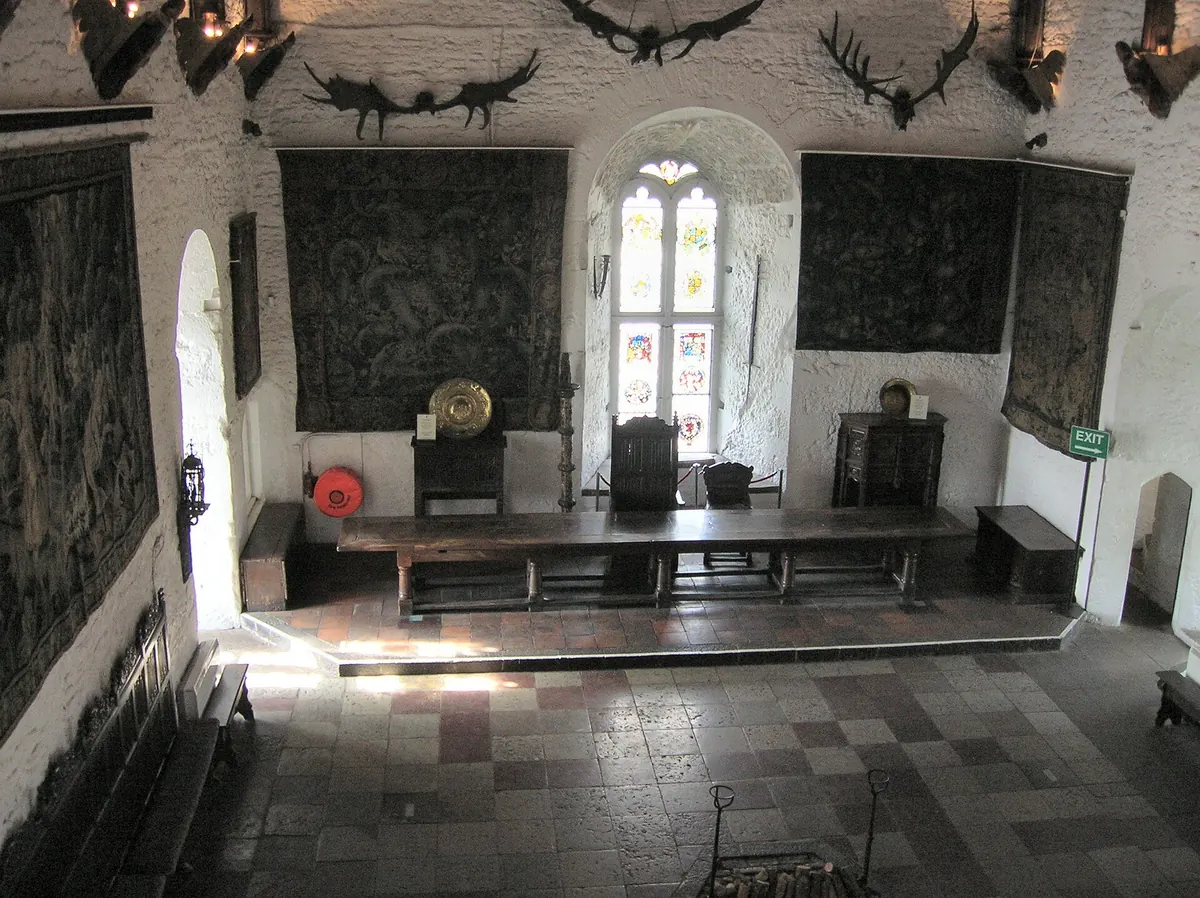 The Great Hall of Bunratty Castle in daylight, a long wooden table on a raised dais beneath tapestries and a stained-glass window, with antlers on the whitewashed walls