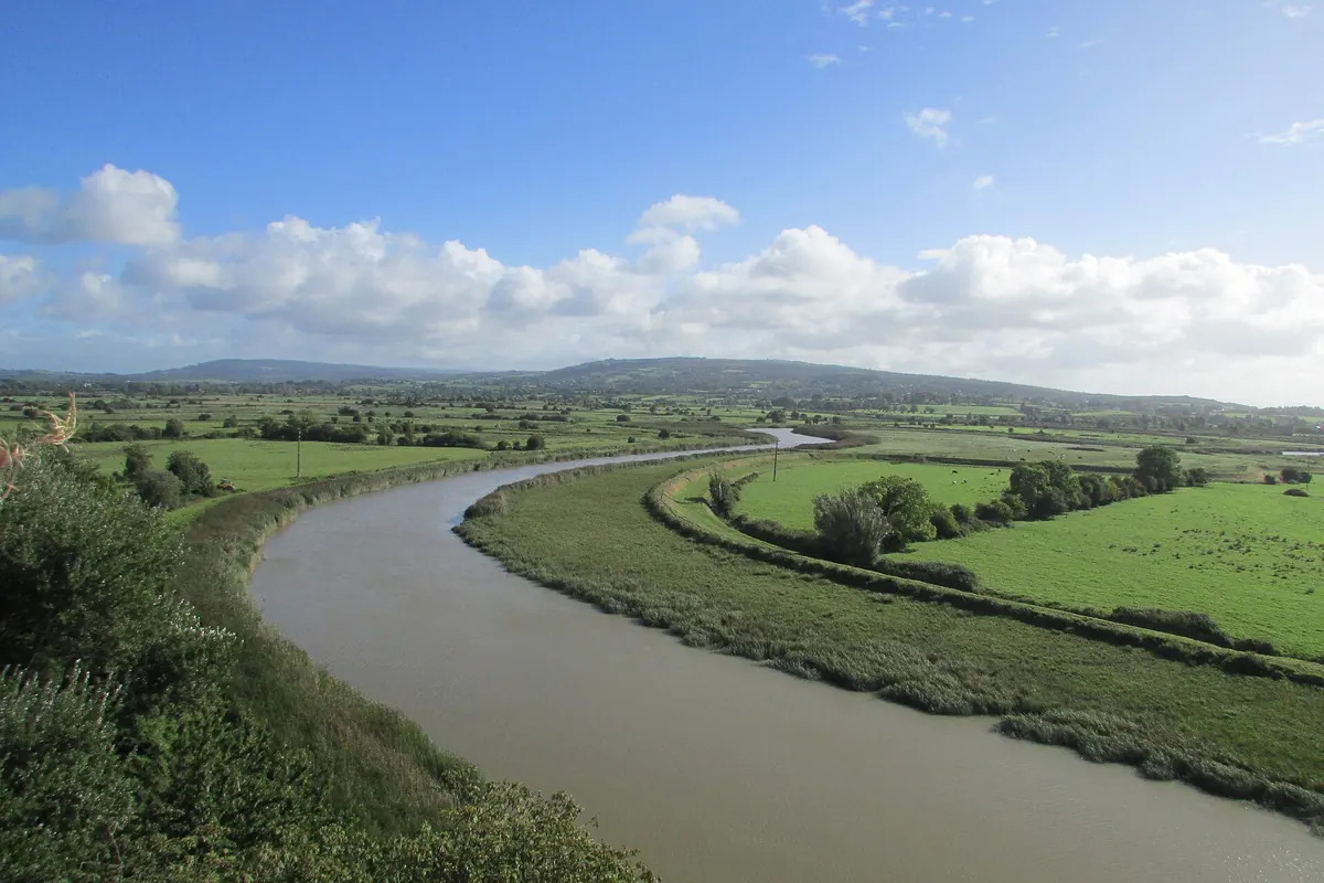 The Ratty river bending through green fields beneath Bunratty Castle, with the Shannon estuary and hills in the distance