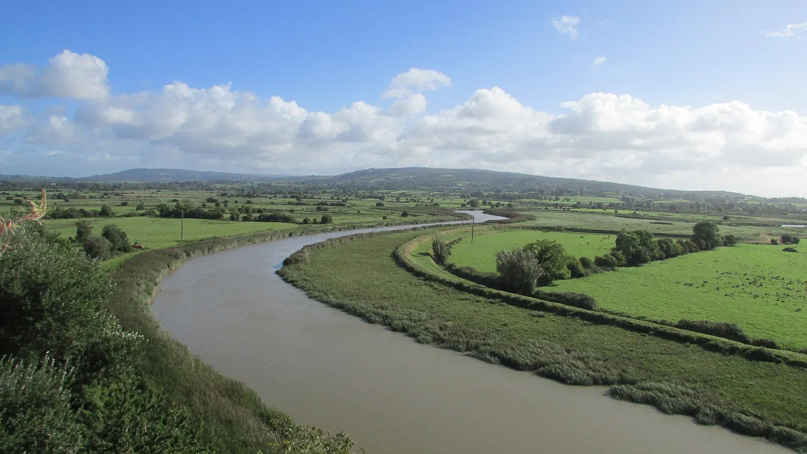 The Ratty river bending through green fields below Bunratty Castle on a bright day, with the Shannon estuary and low hills in the distance
